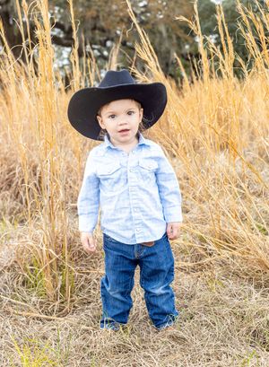 A toddler in a cowboy hat, blue striped shirt and jeans stands in tall golden grass during an autumn photoshoot.