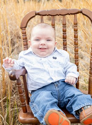 A small child sits in a wooden rocking chair among tall wheat stalks wearing jeans and a light colored shirt.
