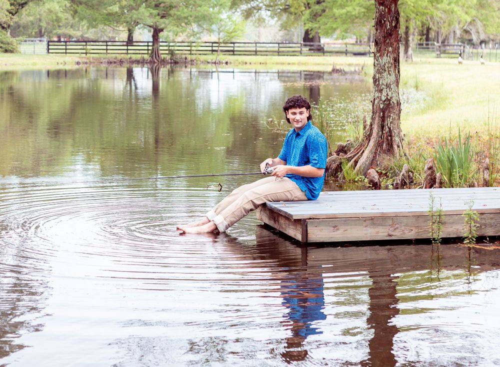 A young person in a blue shirt sits on a wooden dock by a tranquil lake with trees reflecting in the calm water.