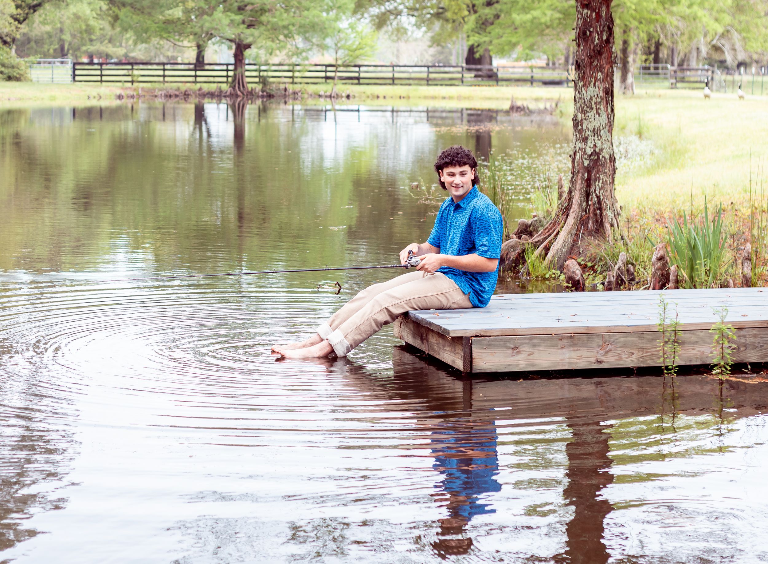 A young person in a blue shirt sits on a wooden dock by a tranquil lake with trees reflecting in the calm water.