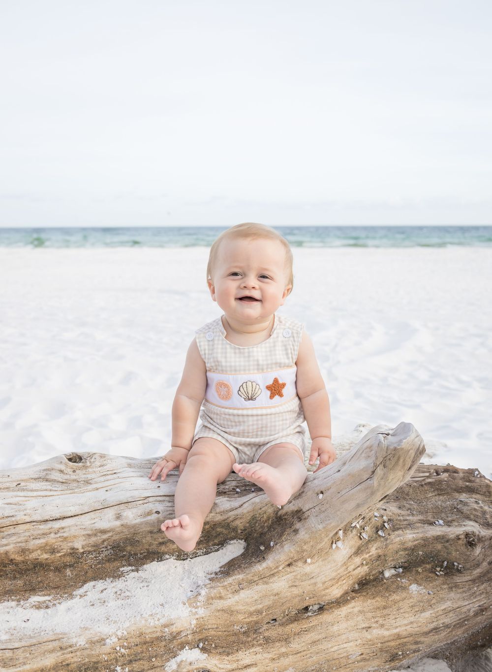 Baby in white outfit sits on driftwood at sandy beach with ocean waves in background.