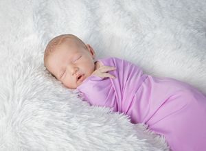 Peaceful newborn baby swaddled in a soft purple wrap, sleeping on a fluffy white blanket.