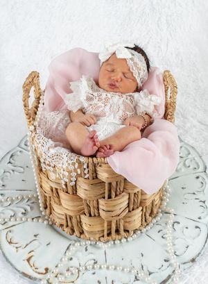 Sleeping newborn baby in lace outfit and bow headband nestled in wicker basket with pink blanket.