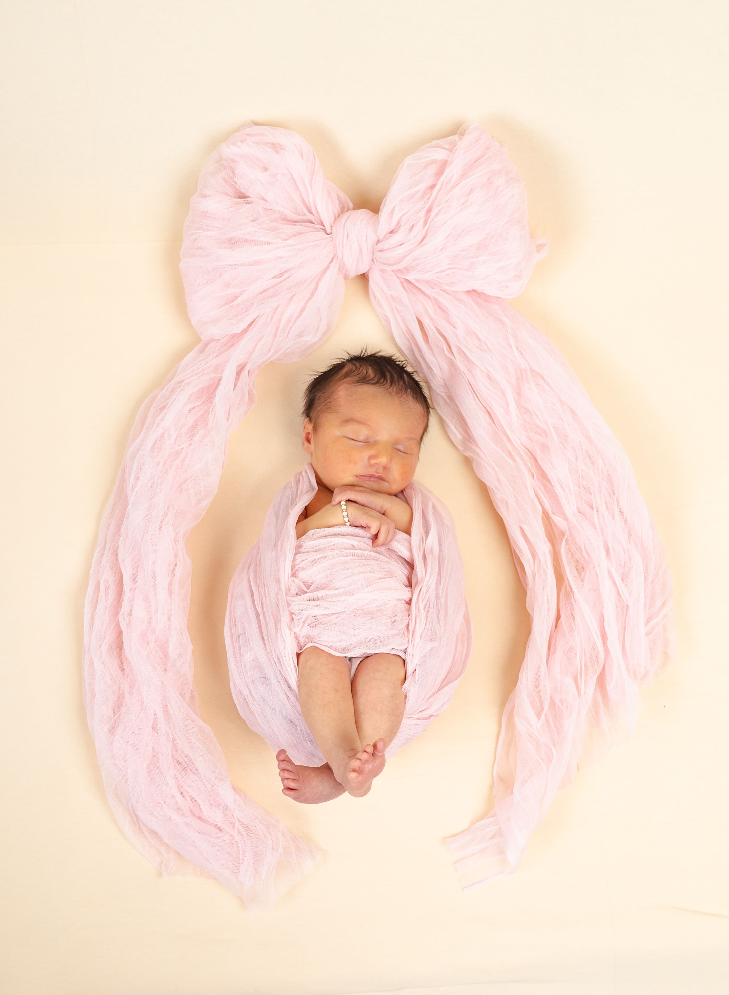 Newborn baby girl swaddled in pink wrap arranged with a large bow on cream background.