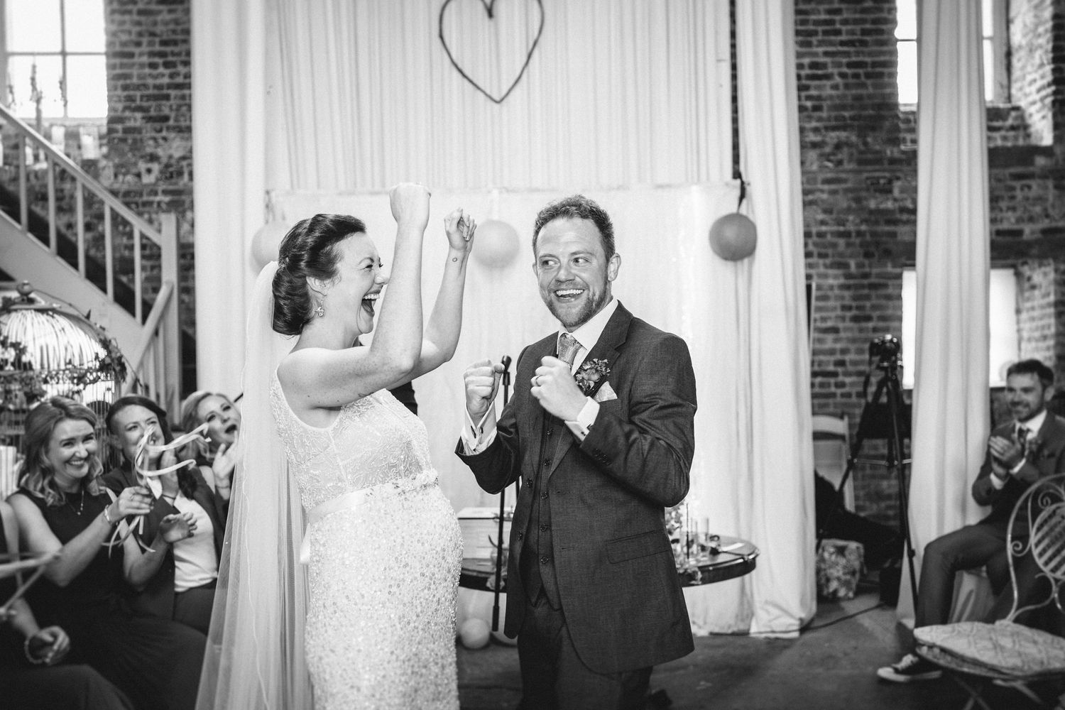 Black and white photo of celebratory moment during indoor wedding ceremony with brick wall backdrop.