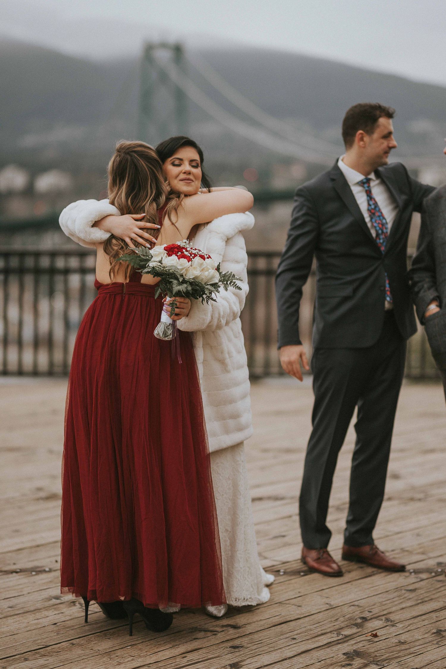 Wedding party embracing on wooden deck with bridge view in background.
