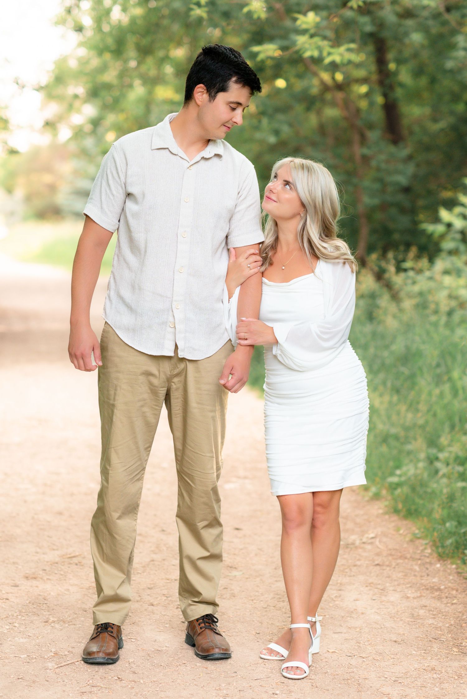 A couple in casual attire walks together on a dirt path surrounded by green foliage on a sunny day.