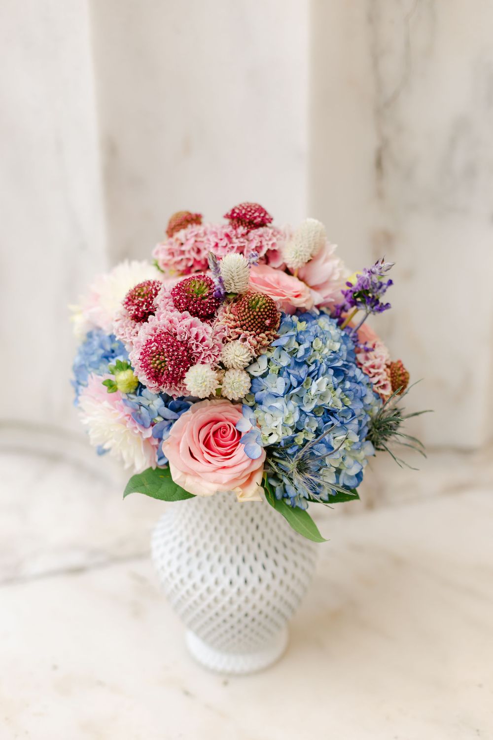 Colorful wedding bouquet with pink roses, blue hydrangeas in a white hobnail vase.
