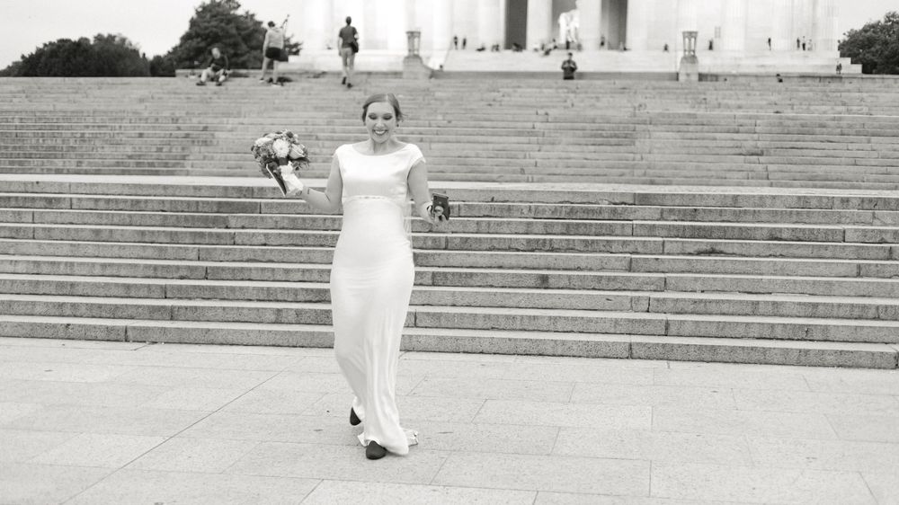 Bride in elegant white gown holds bouquet on steps of Lincoln Memorial in black and white photo.