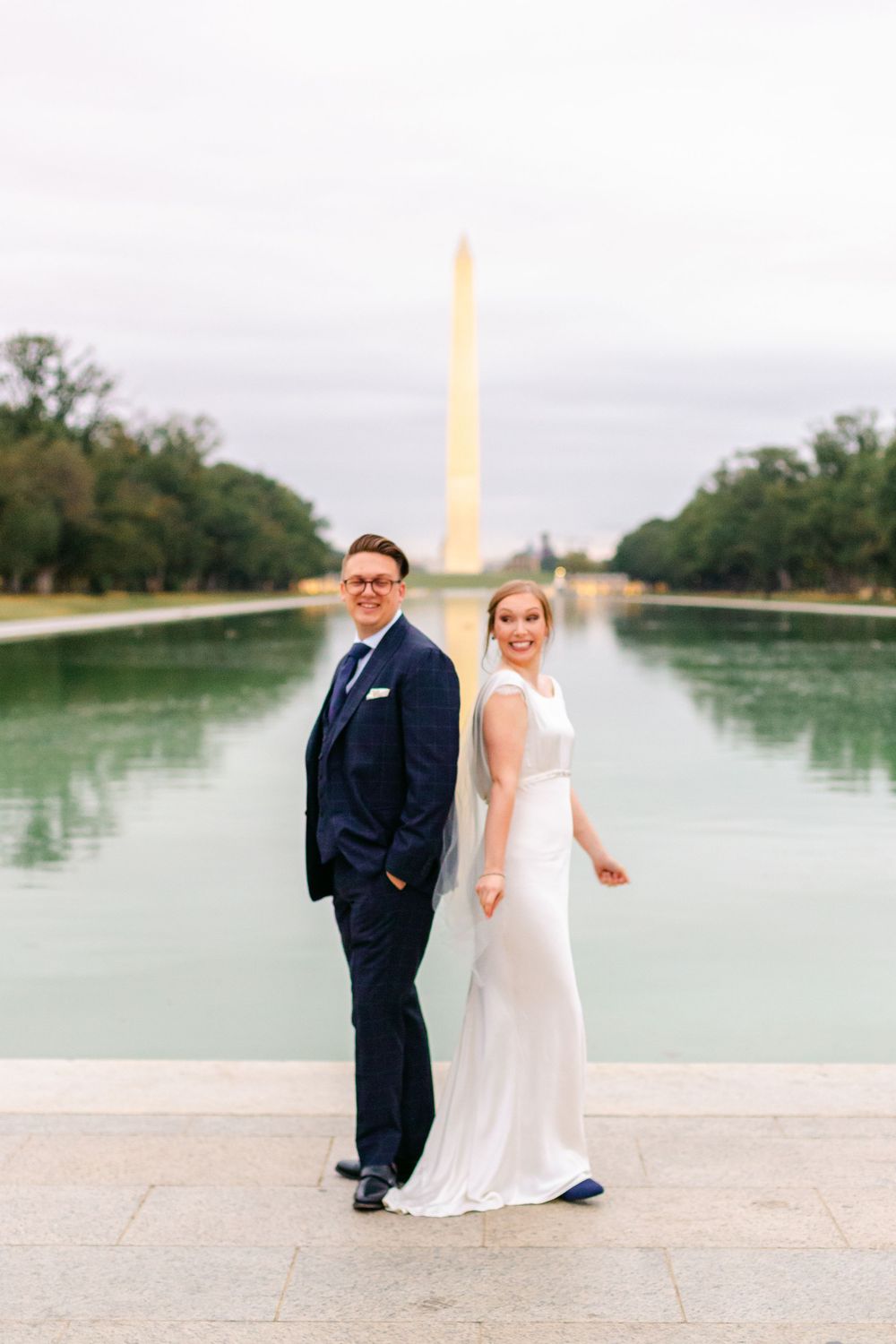Bride and groom smile at camera with Washington Monument reflected in the pool behind them.