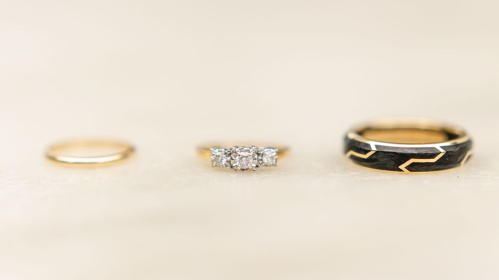 Three wedding rings displayed on neutral background: gold band, diamond engagement ring, and black ring.