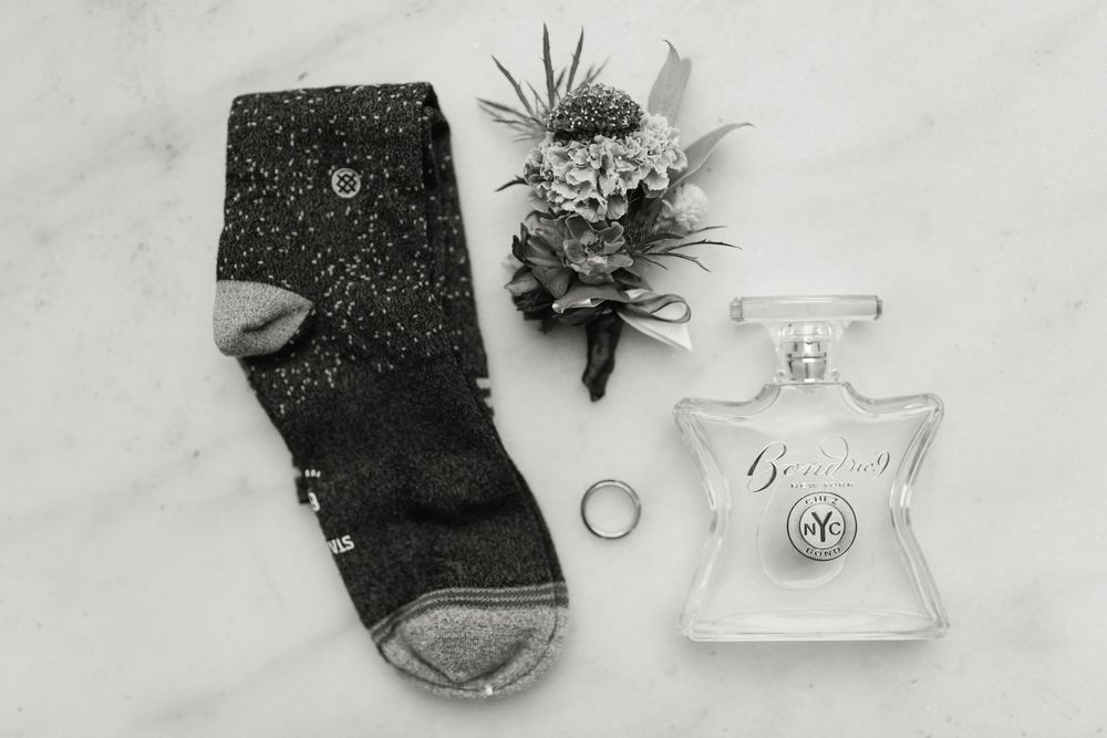 Black and white flat lay of groom accessories including socks, boutonniere, and cologne.