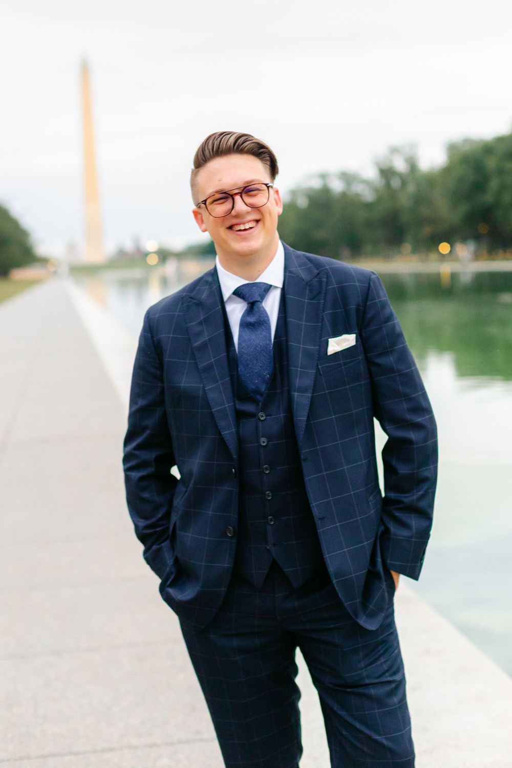 Smiling groom in navy plaid suit standing beside the Reflecting Pool with Washington Monument.
