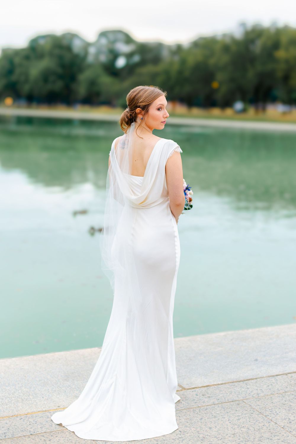 Bride in elegant white backless gown with draped detail stands by reflecting pool.