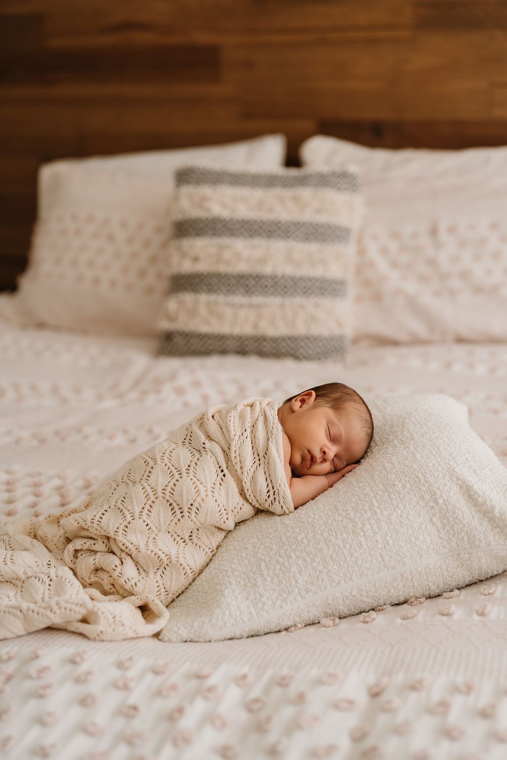 A sleeping newborn baby wrapped in a cream-colored knit blanket on white textured bedding.