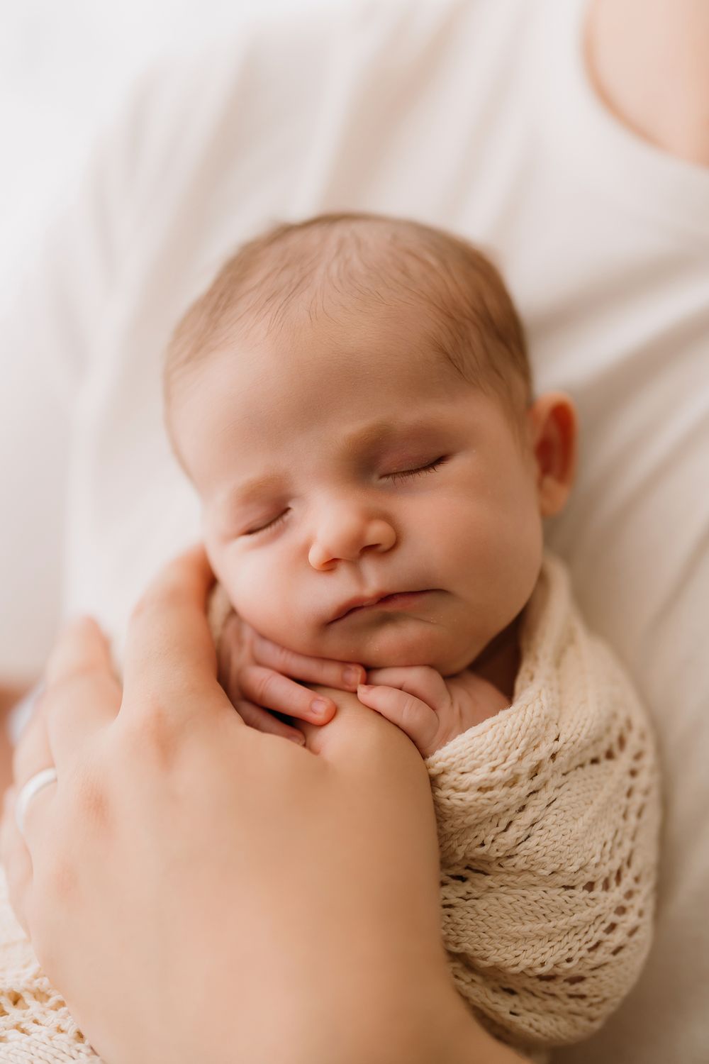 A sleeping newborn baby wrapped in a beige knit blanket lies peacefully against a soft cream background. Sydney lifestyle newborn photograph