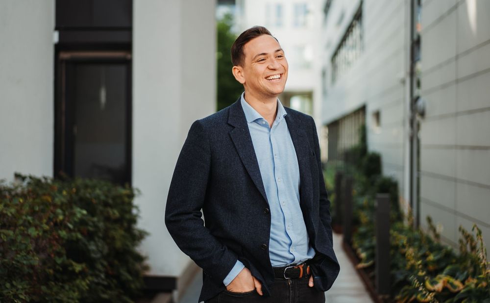 Professional in navy suit jacket and blue shirt stands outdoors by modern building.