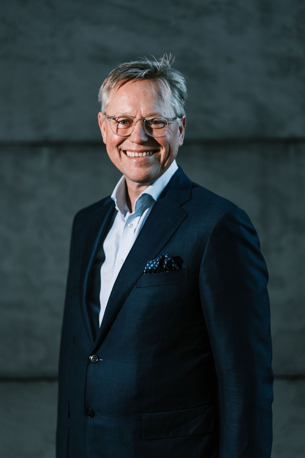 Professional wearing navy suit and glasses against industrial backdrop.