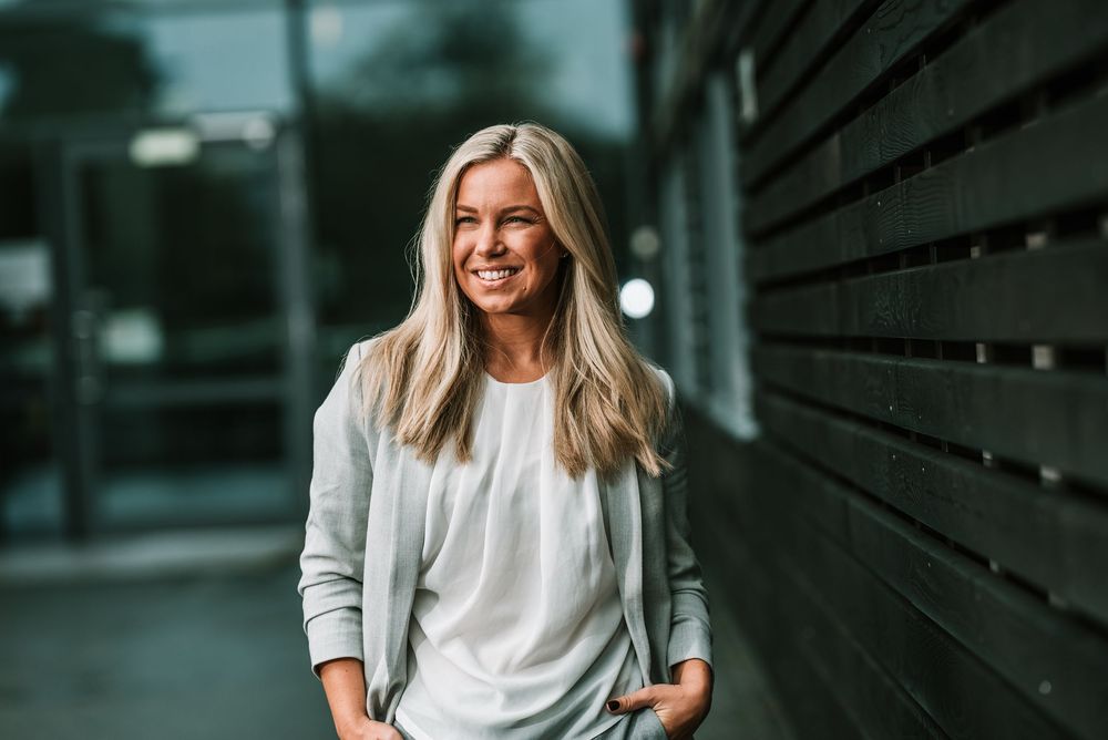 Professional in casual gray blazer and white top poses against dark wall in urban setting.