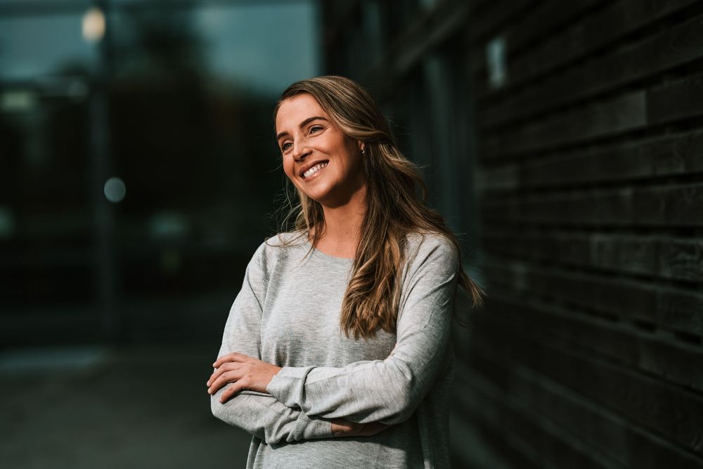 Person in gray sweater leans against dark wall while smiling.