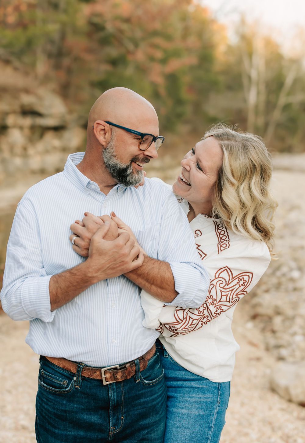 A loving couple shares a tender moment outdoors during an autumn photoshoot, both wearing casual attire and sharing genuine laughter.
