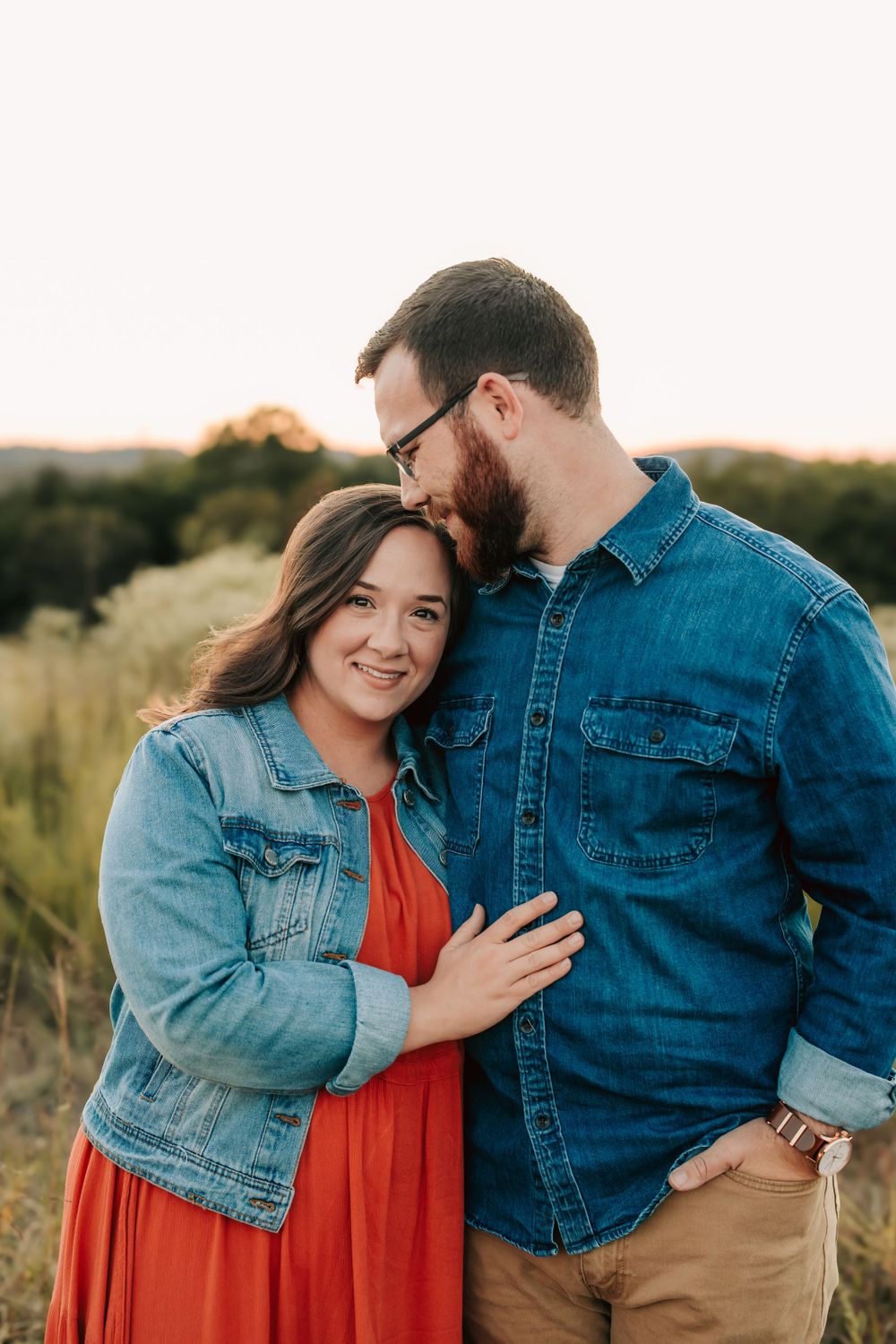 A couple wearing denim jackets embrace in a sunset field with orange glow and tall grass in background.