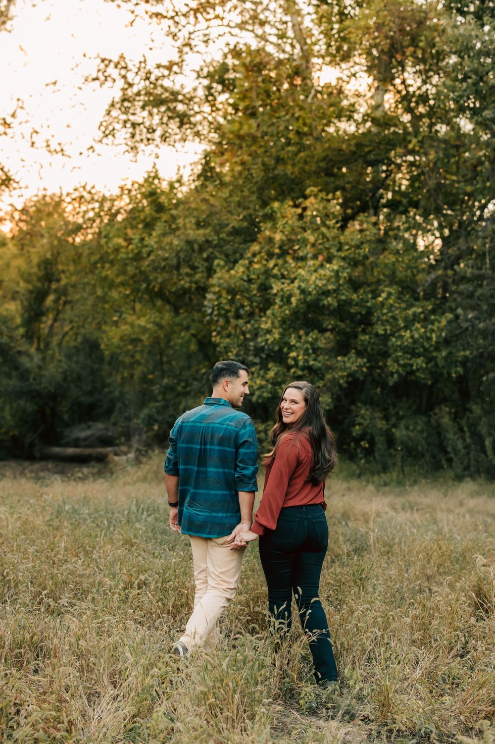 A couple walks through a sunlit meadow surrounded by autumn trees at golden hour.