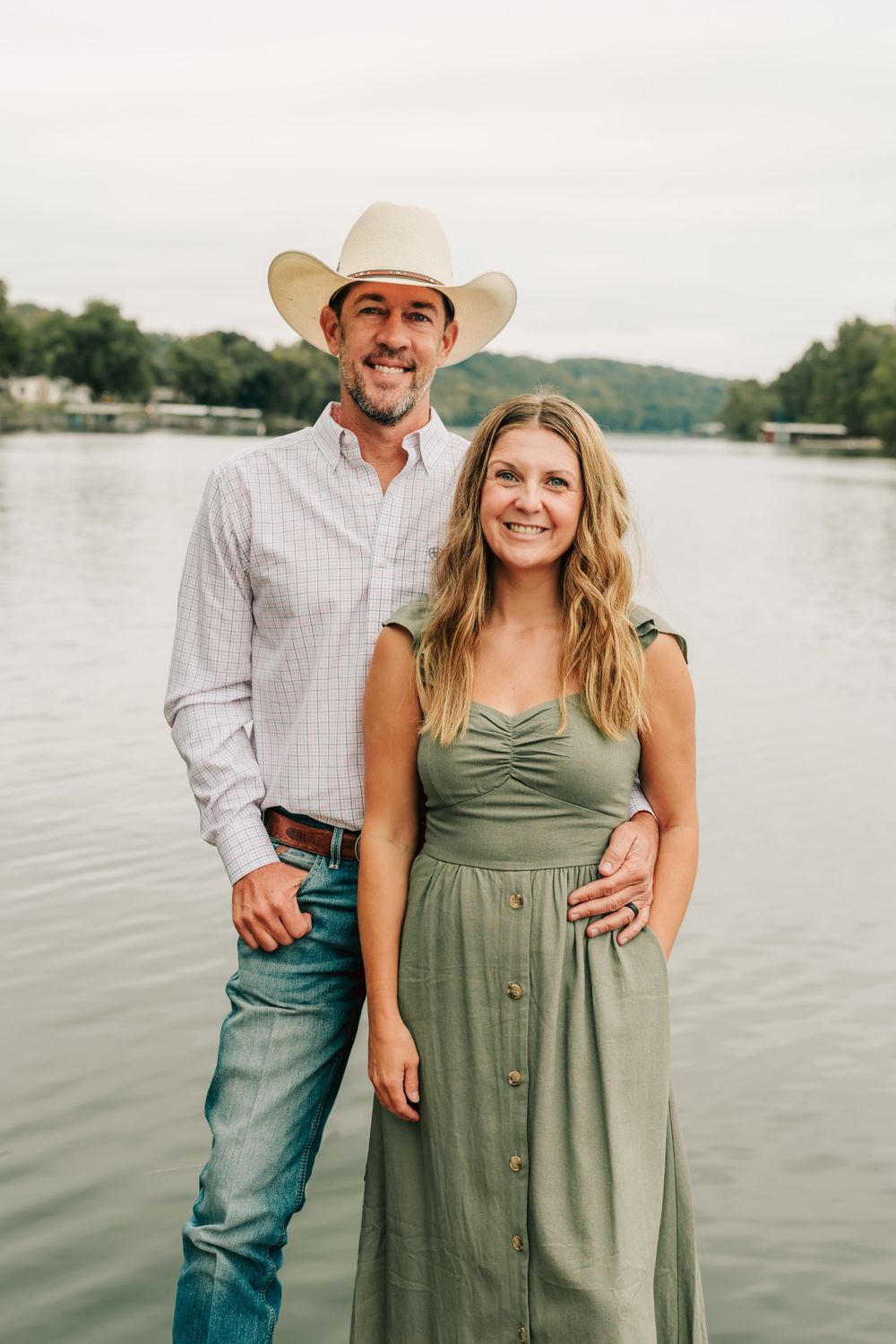 A couple poses together by a lake, the person wearing a cowboy hat and button-down shirt with someone in a sage green dress.