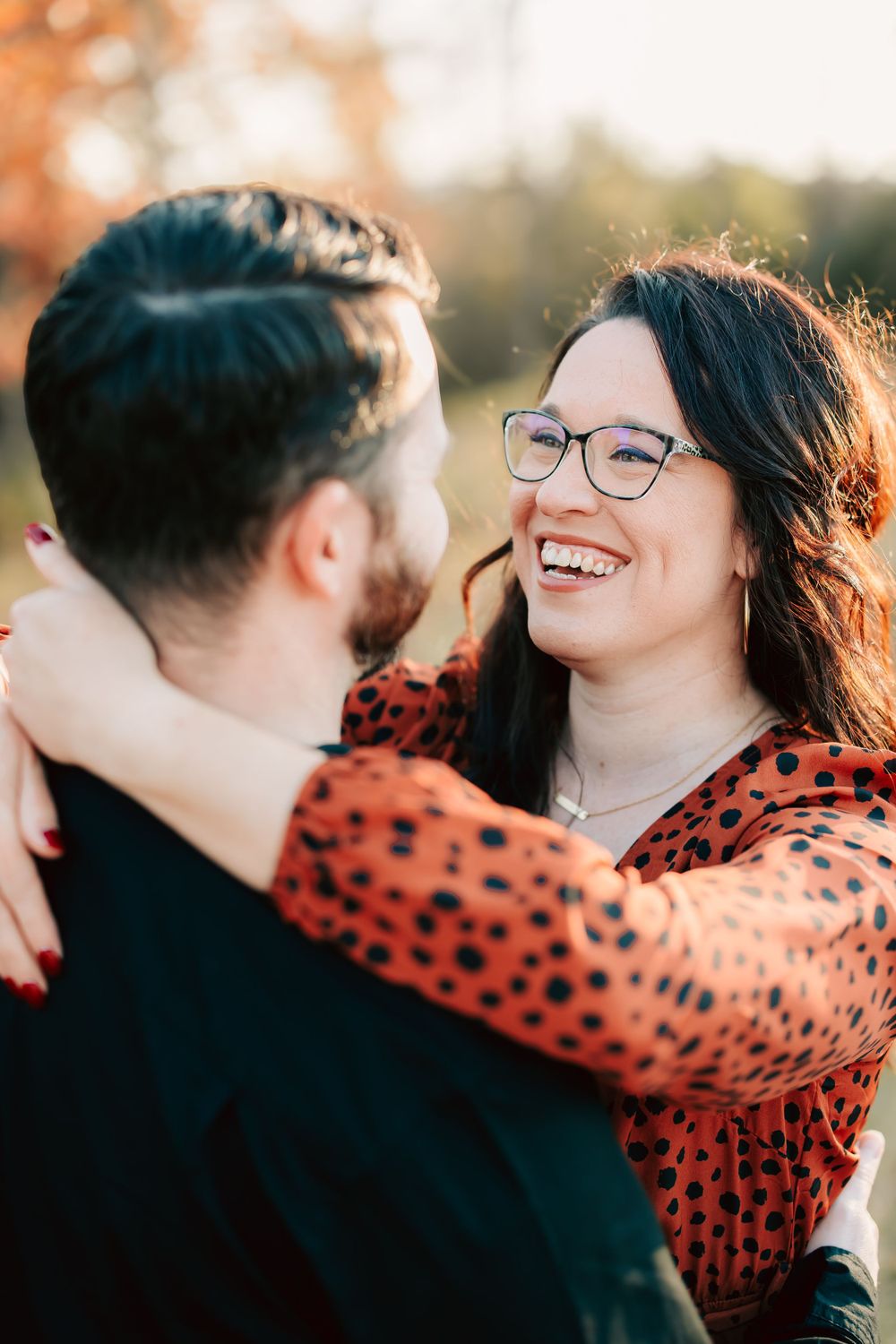 A joyful couple embraces outdoors during golden hour, with a person in glasses wearing a red polka dot top.