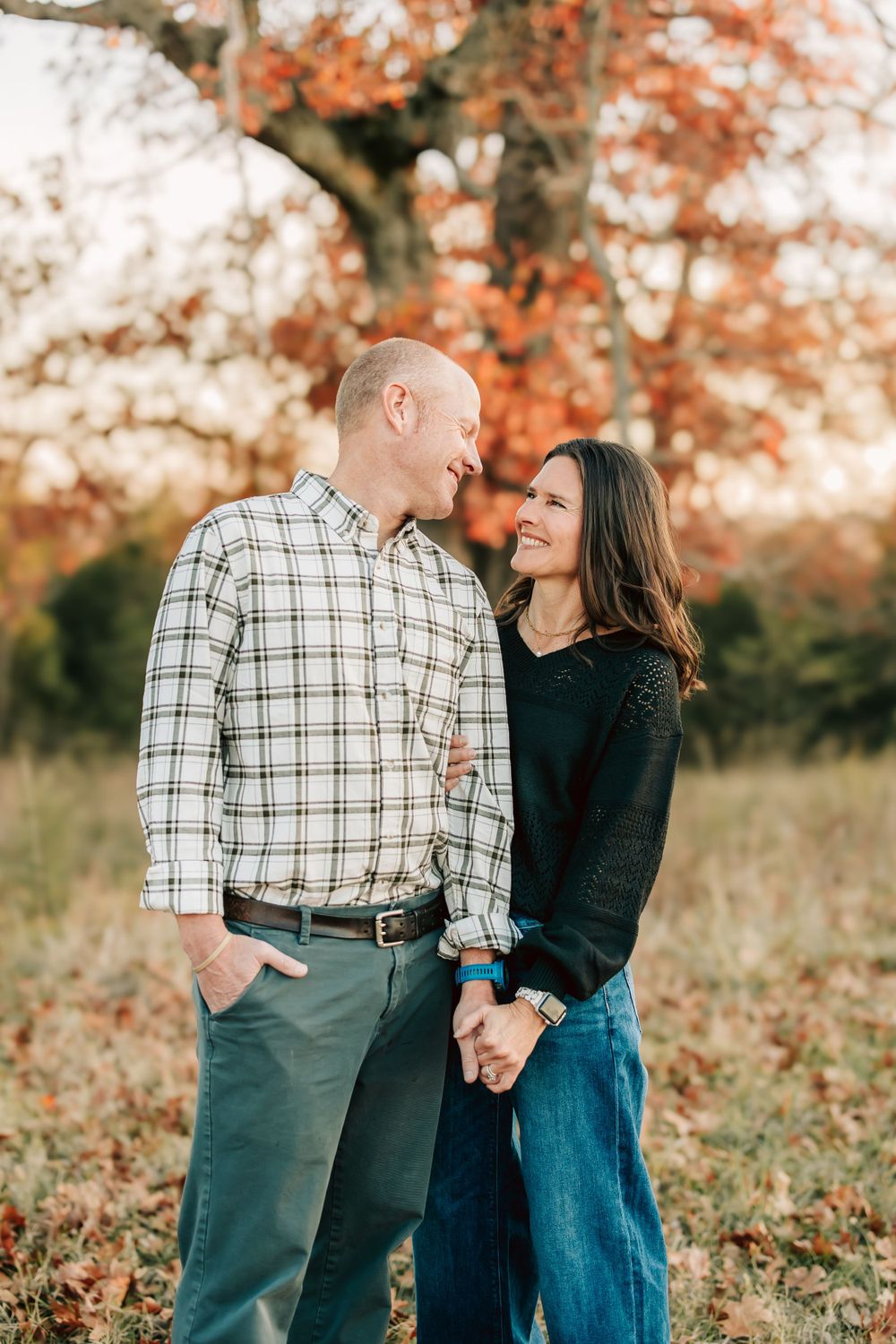 A couple shares a loving moment while holding hands in an autumn setting with orange foliage in the background.