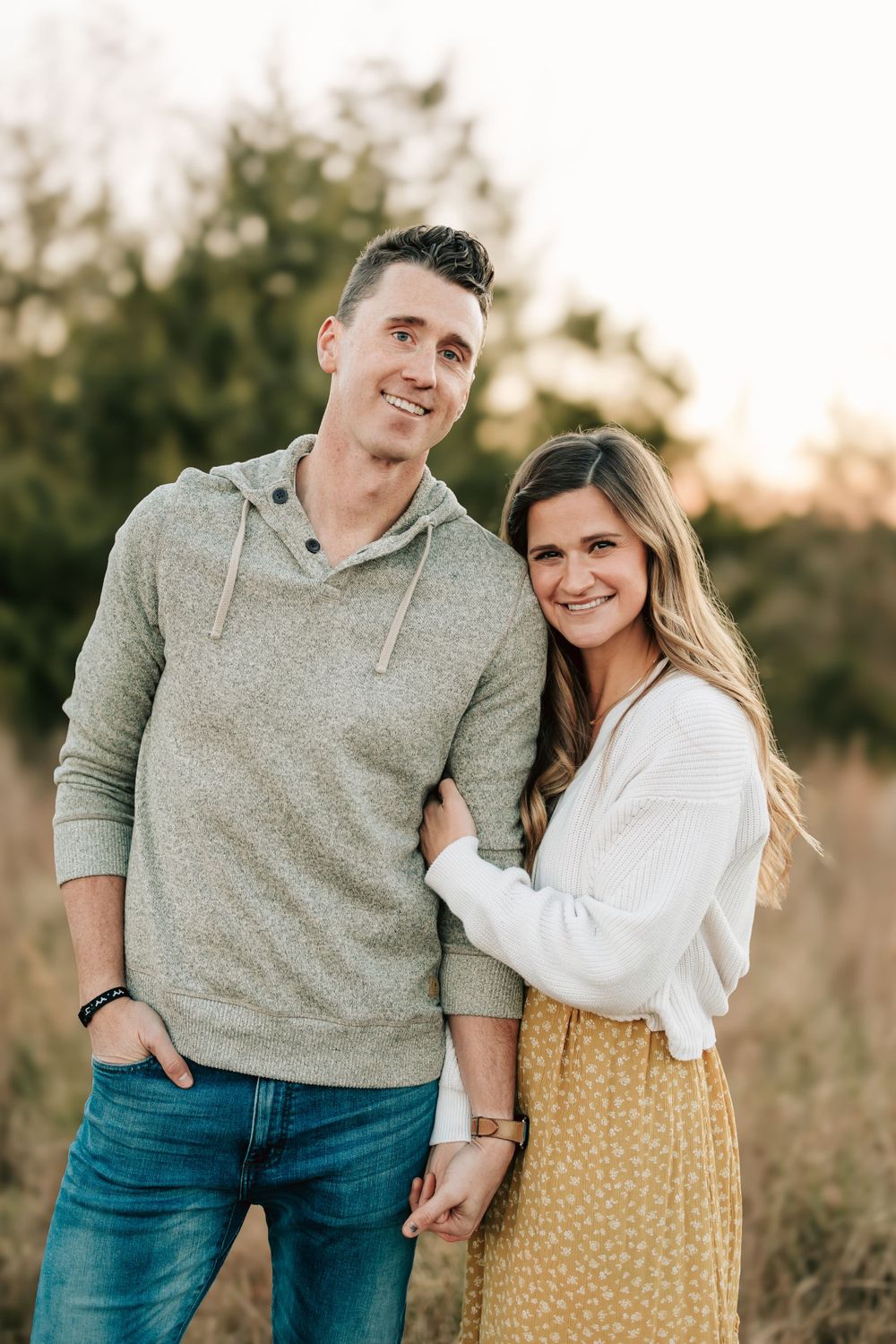 A couple poses together in a field at sunset wearing casual fall attire including a hoodie and floral skirt.
