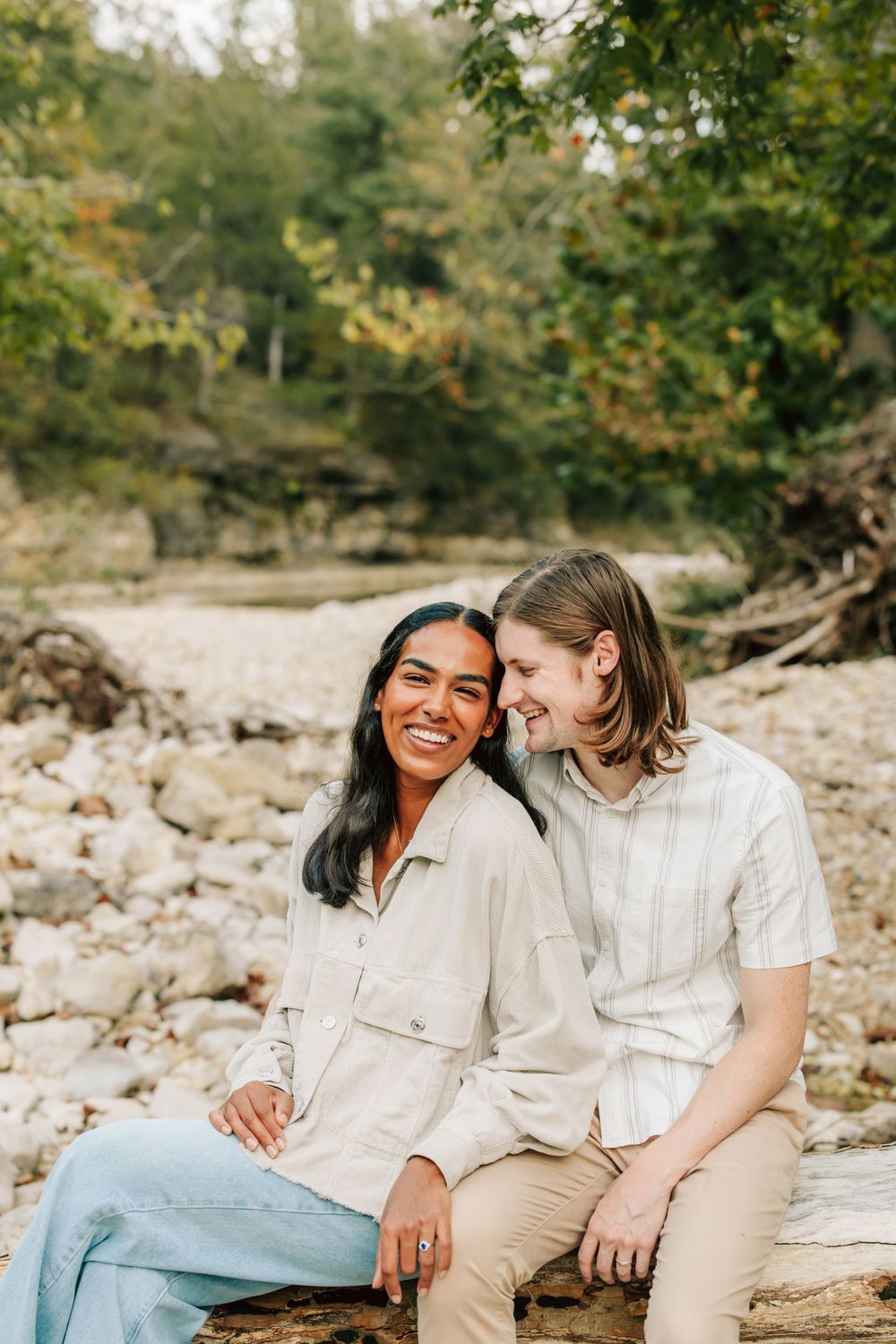 A couple shares a joyful moment together while sitting on rocks in a natural outdoor setting during autumn.