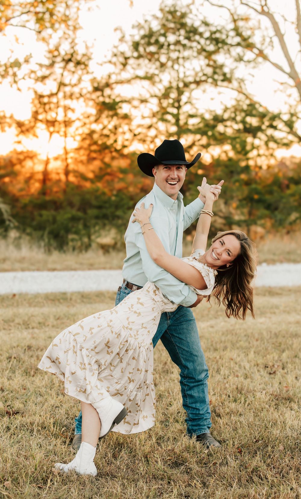 Couple dances together in a field at sunset with cowboy hat and floral dress creating a romantic country scene.