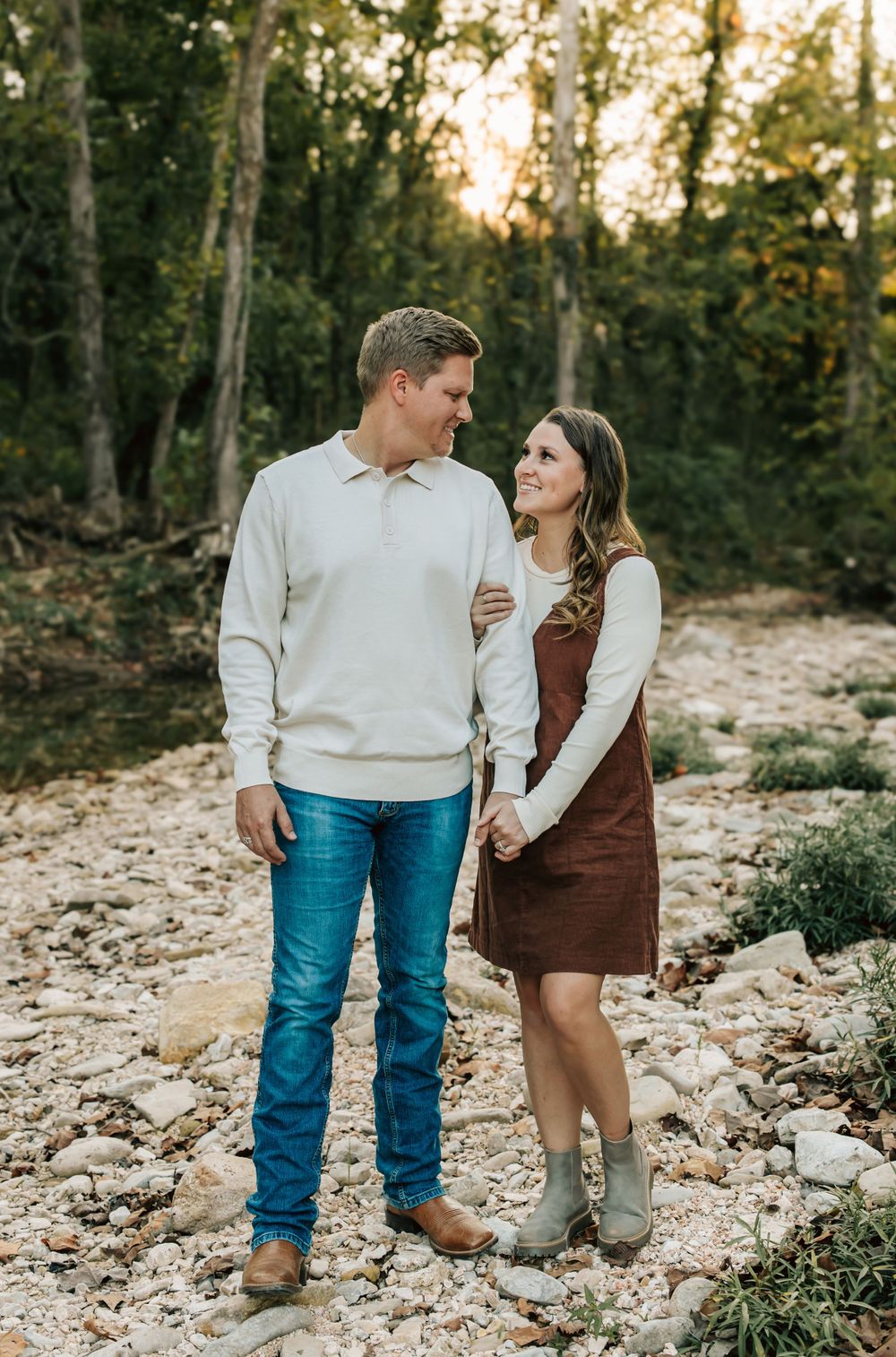 A couple in casual attire shares a romantic moment on a rocky path in the woods during sunset.