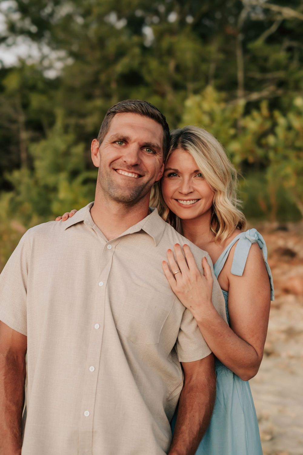 A couple smiles warmly together outdoors during a sunset photoshoot, wearing casual summer attire.