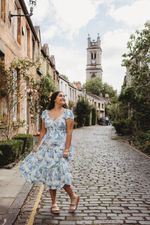 A blue floral dress catches the breeze on a cobblestone street with a historic church tower in the background.