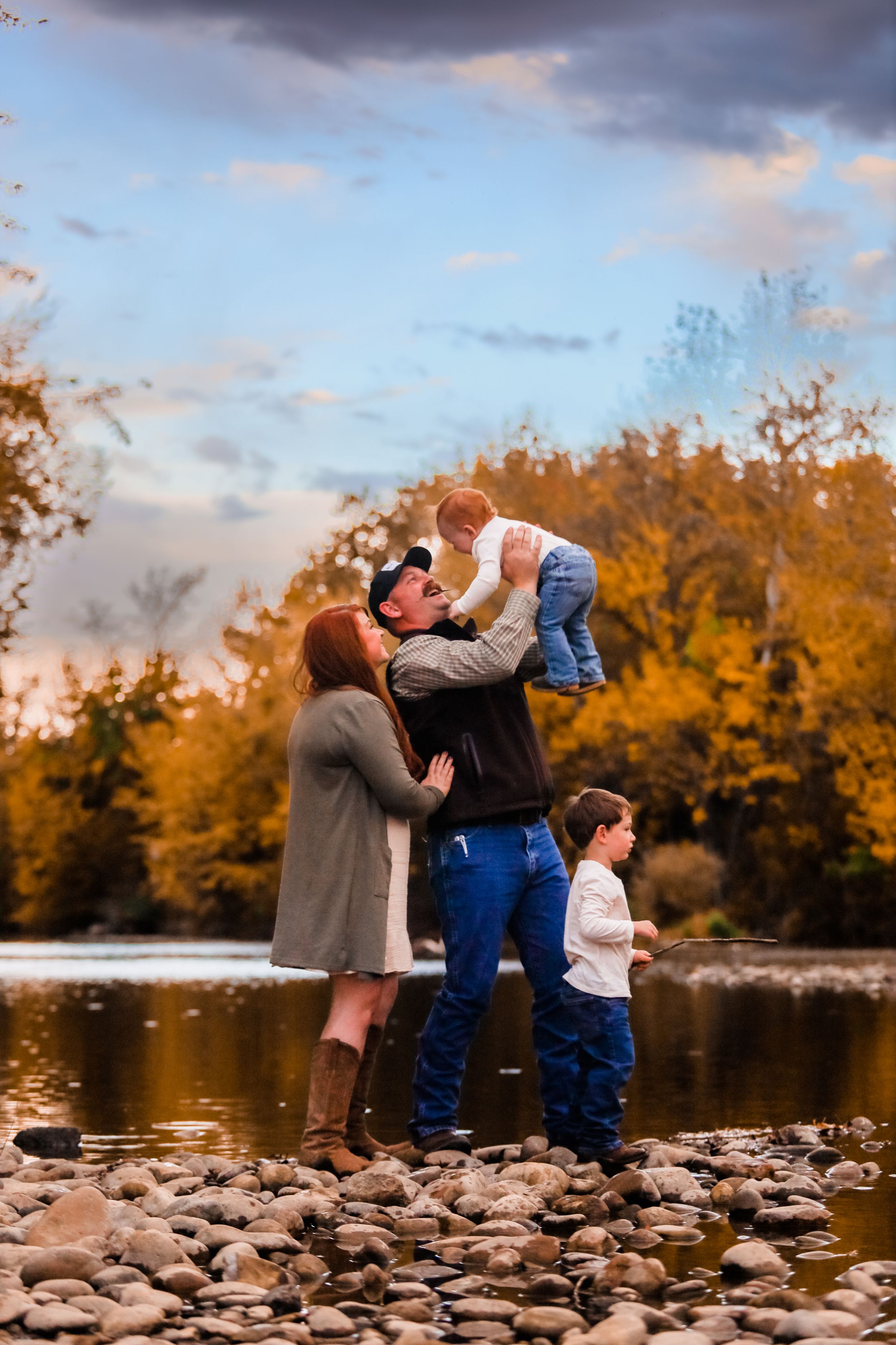 People gather on rocky riverbank with autumn trees and cloudy sky in background.