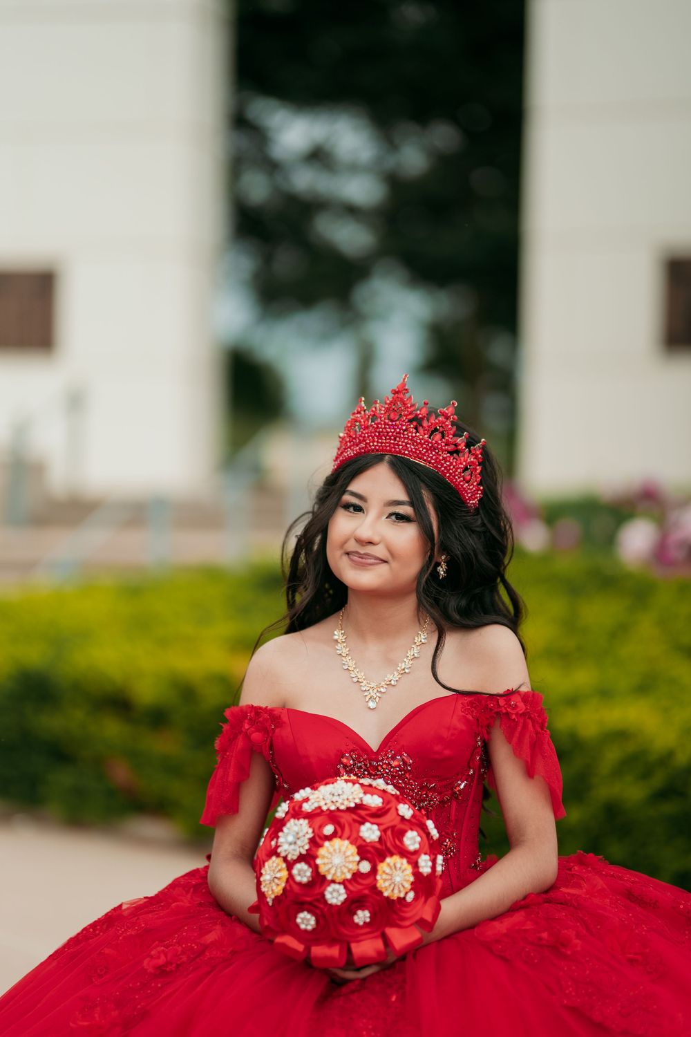 Red quinceañera gown with off-shoulder design and crystal embellishments in garden setting.