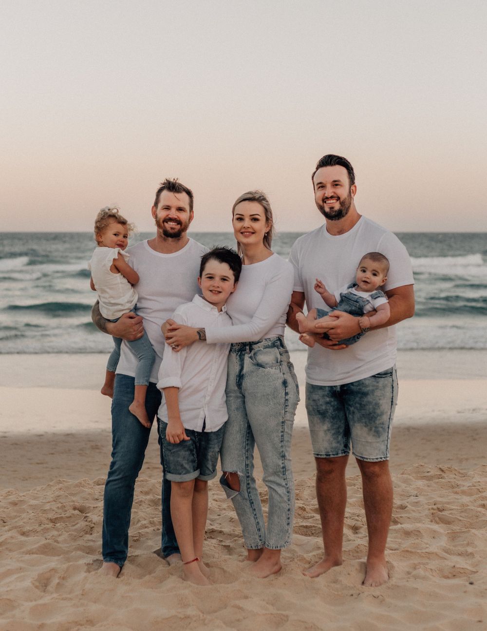Family group poses casually on beach at sunset wearing coordinated white and denim outfits.