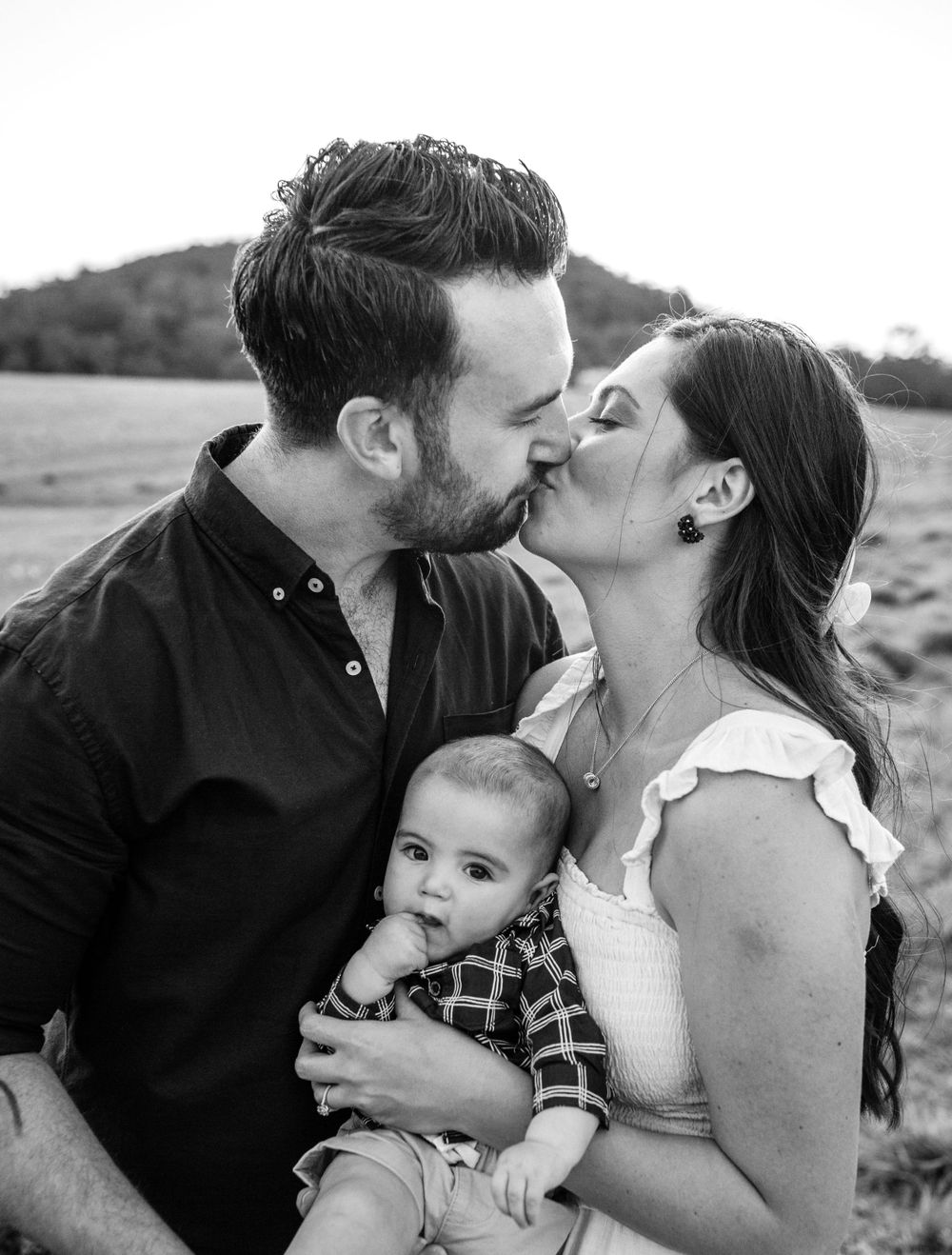 Family portrait outdoors with baby in plaid shirt against natural landscape.
