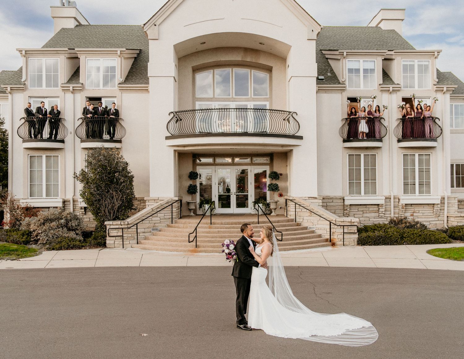 A couple standing outside an elegant French-style mansion with curved balconies and grand stone staircase.