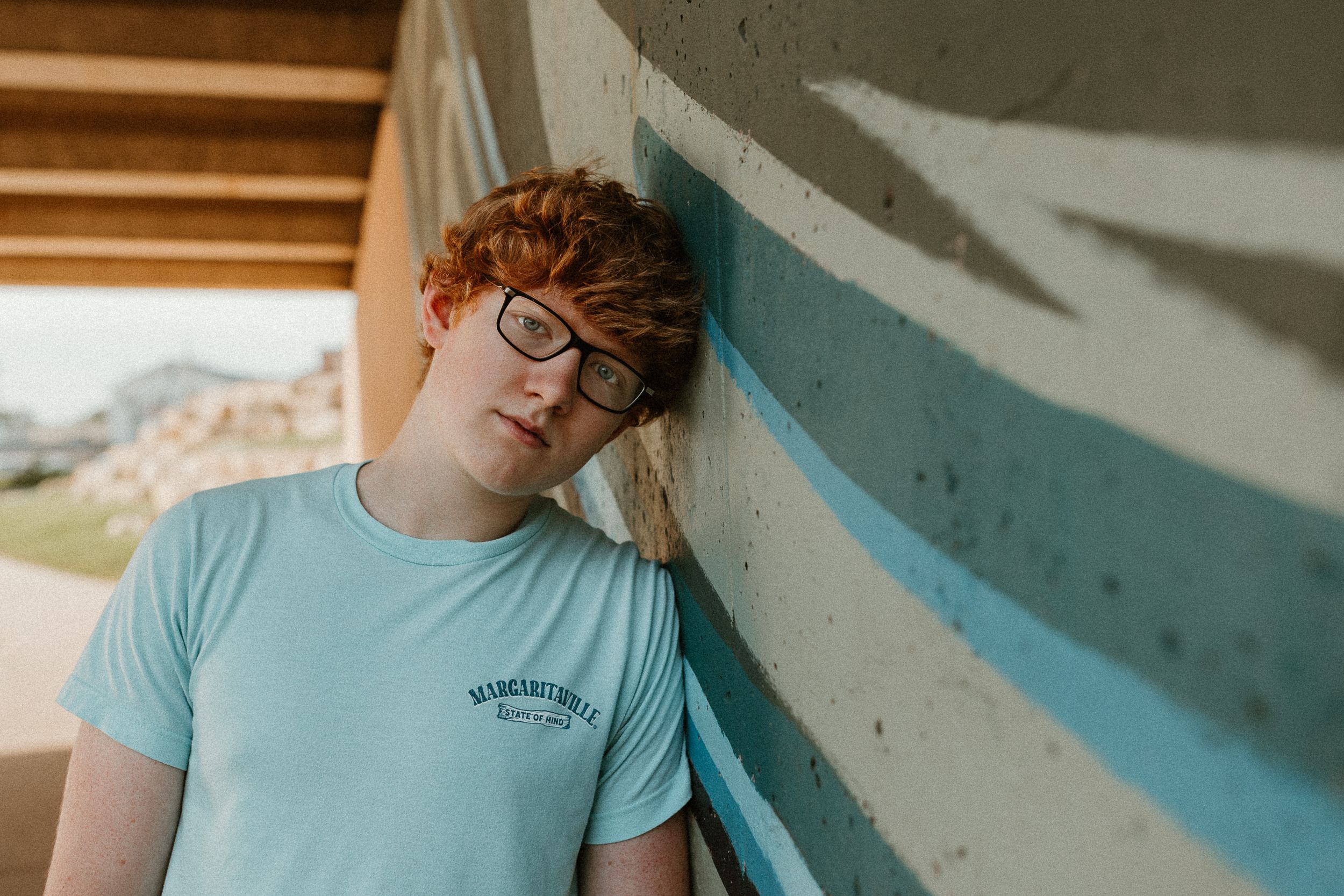 A person in glasses and light blue t-shirt leans against a wall with painted stripes.