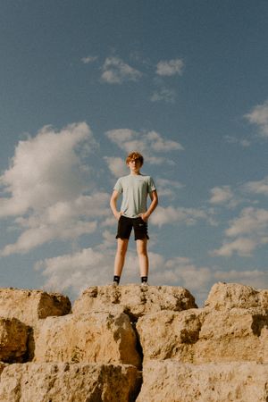 A figure stands atop rocky terrain against a blue sky with white clouds.