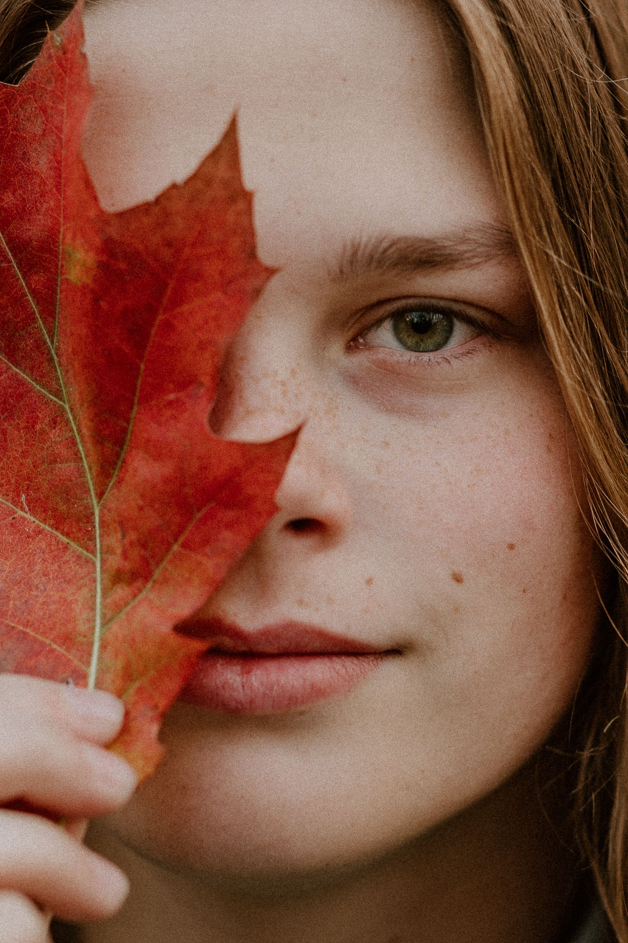 Creative portrait with a red maple leaf covering half the face against neutral background.