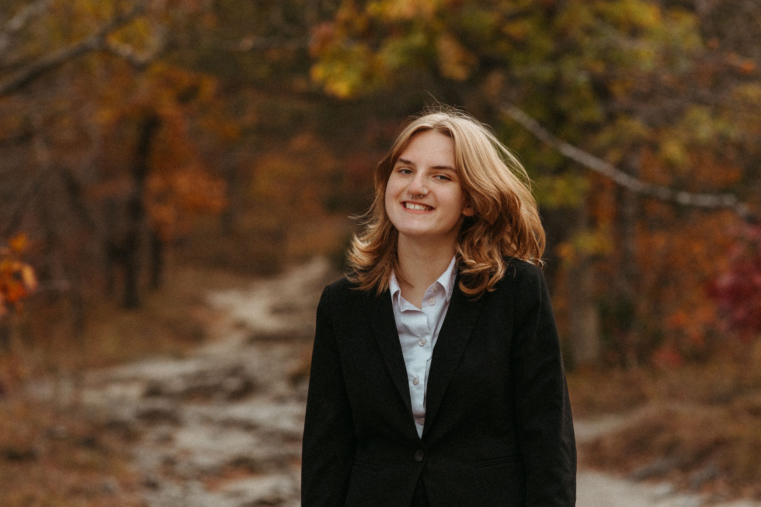 Person wearing black coat smiles with windswept hair against autumn foliage background.