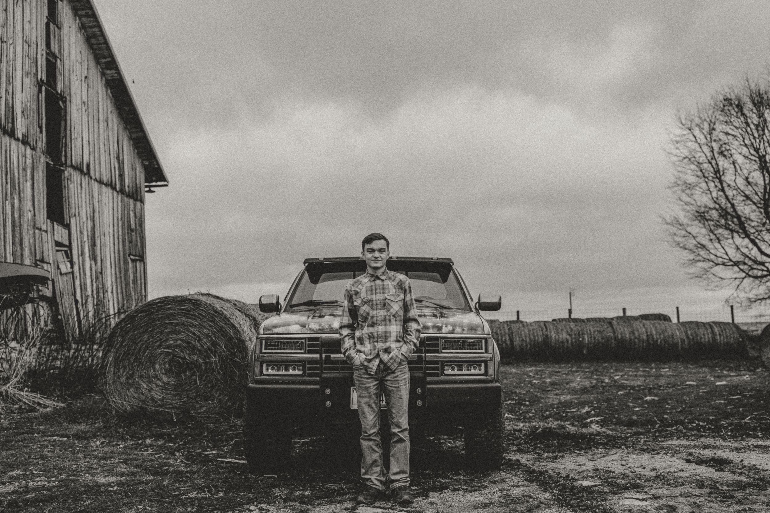 Black and white photo of someone standing in front of a pickup truck near a barn.