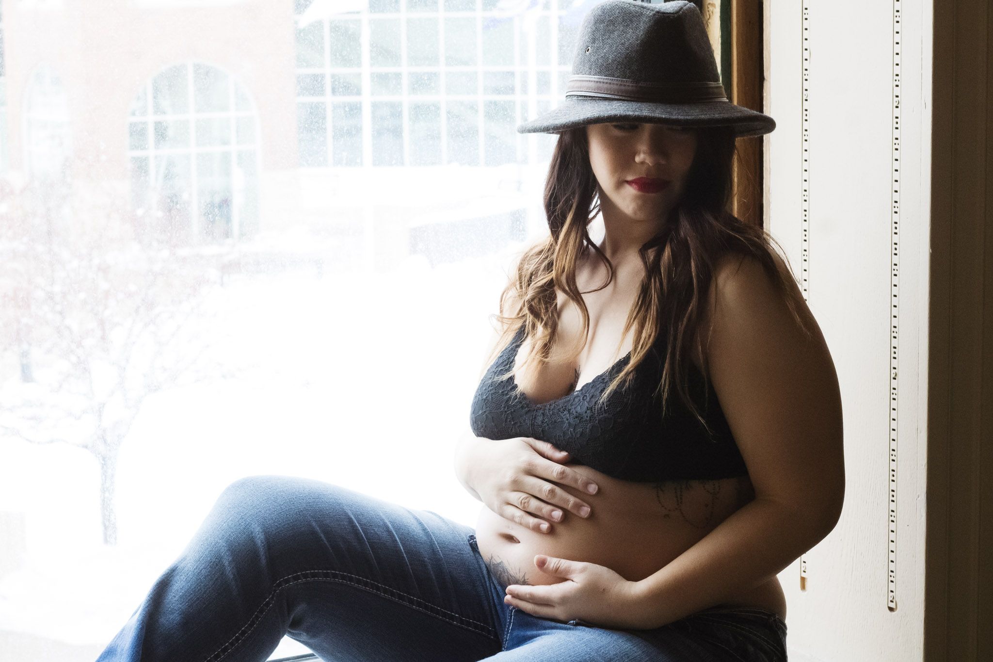 Person in black top and hat sits by window during intimate photo session.
