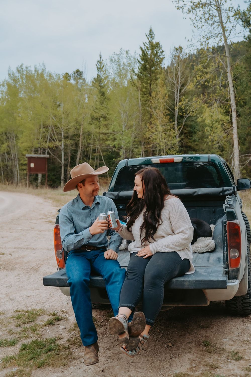 A couple sit together on a pickup truck tailgate having drinks in a wooded area on a casual afternoon.