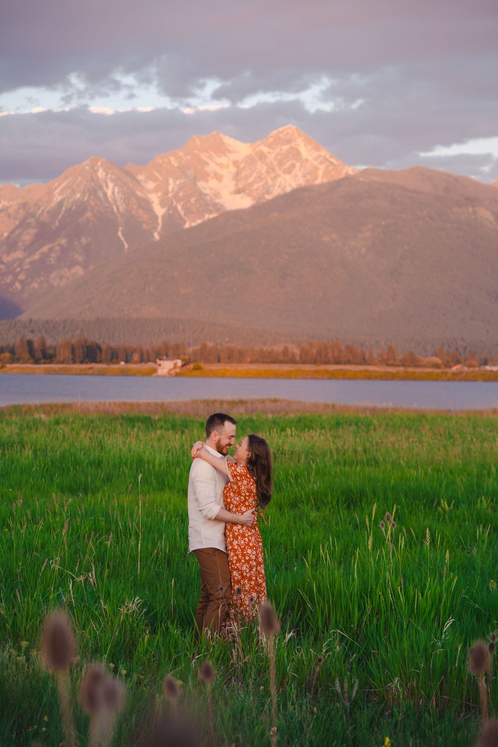 A couple embraces in tall grass at sunset with snow-capped mountain peaks and a lake in the background.
