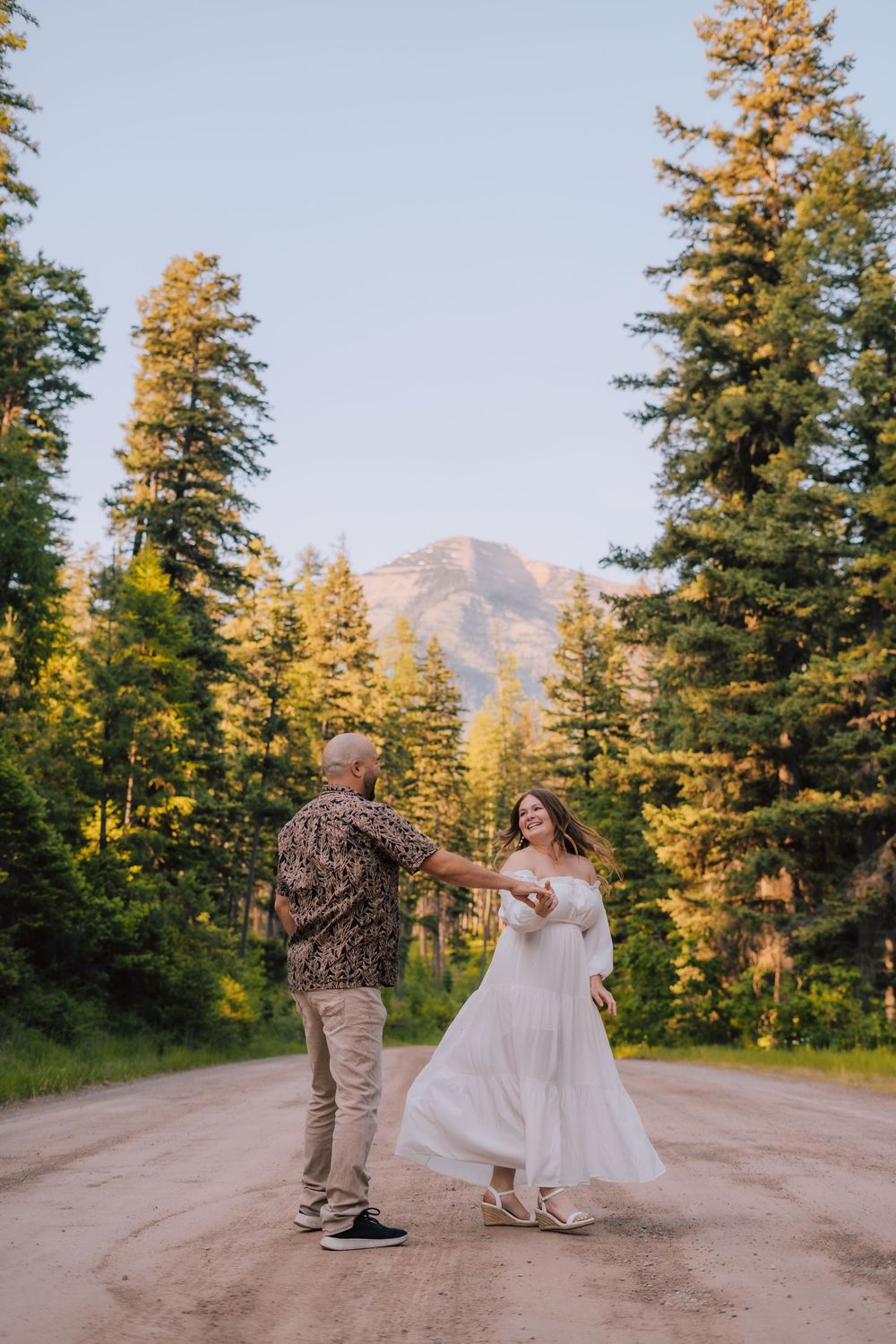 Couple in wedding attire stands in a mountain forest setting during sunset with golden light filtering through trees.